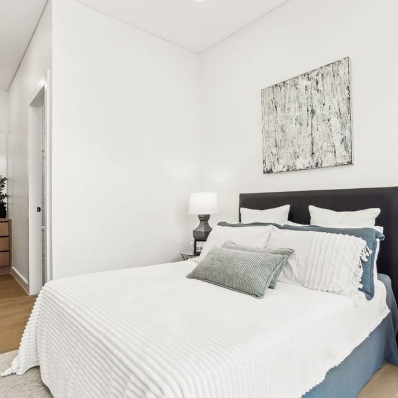 Cosy bedroom at 55 Eyre Street, Chifley, 2036-Duplex featuring a plush bed with white and blue bedding, a bedside lamp, and abstract wall art in a bright, minimalist space.