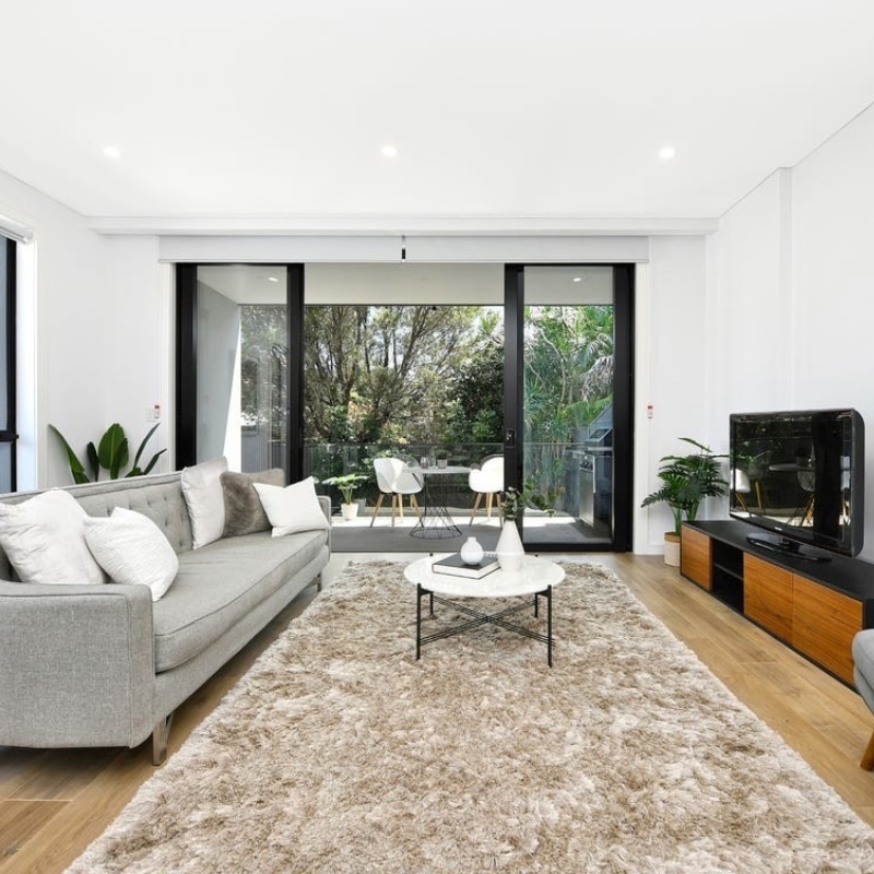Living room with grey couch and cushions, extending to a stylish outdoor coffee table area through a glass door at 91 King Street, Mascot.