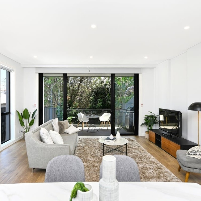 Living room with a white couch and pillows, opening to an outdoor coffee area at 91 King Street, Mascot — Sydney East Building & Renovations project.