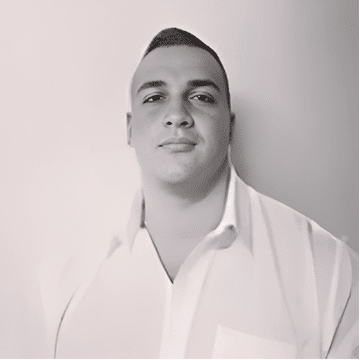 A person wearing a white shirt stands against a light-colored wall, presenting a neutral expression.