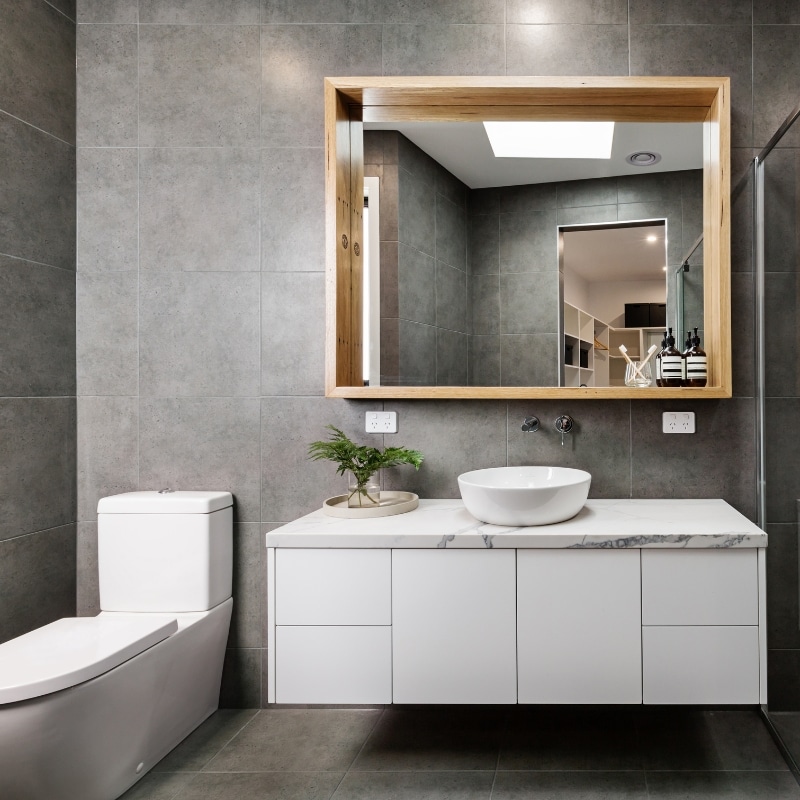 Bathroom Renovation – Modern bathroom with wall-hung toilet, sleek vanity with round sink, and large wooden-framed mirror against grey tiled walls