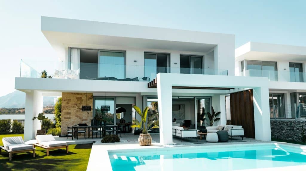 Modern double-storey luxury home with large glass windows, outdoor lounge, swimming pool, and landscaped garden under a clear blue sky.