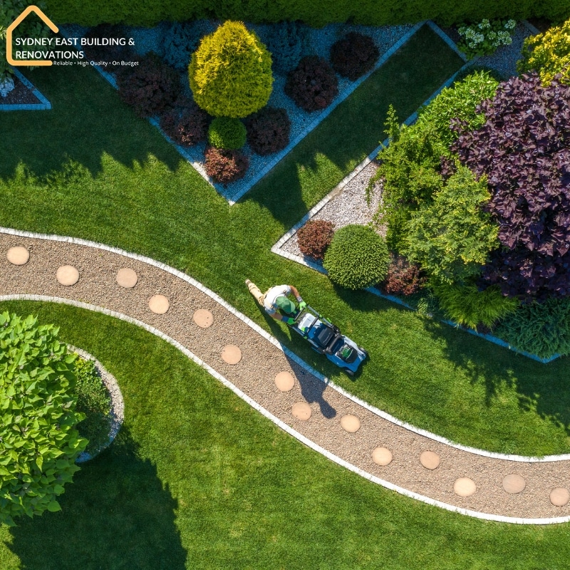 Aerial view of expert landscape design and construction featuring a gardener mowing a lush lawn with shrubs and decorative stone pathways.