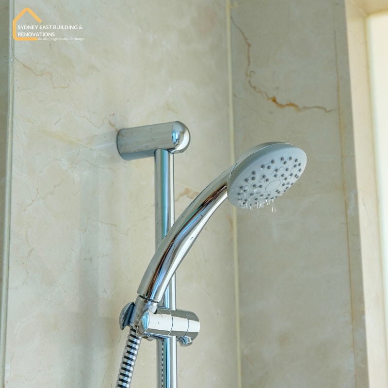 Shower leak repairs shown with water dripping from a handheld showerhead in a marble-tiled bathroom.
